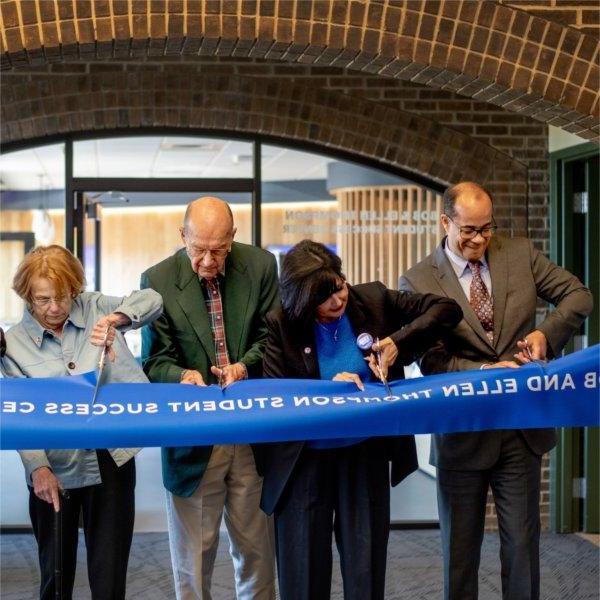 people stand behind a blue ribbon, Bob and Ellen Thompson Student Success Center, with scissors in hand to cut the ribbon